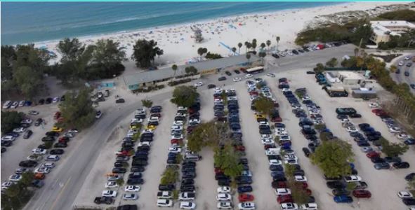 An aerial photograph of a crowded beach parking lot, showing rows of cars parked near the shore of a blue ocean.